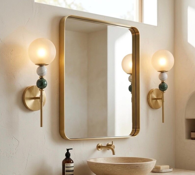 May include: Bathroom wall sconces with a gold finish, featuring a frosted glass globe, and decorative marble and green stone accents. A gold-framed mirror hangs above a stone sink. The sconces are approximately 12 inches tall.