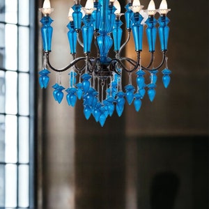 May include: A vibrant blue chandelier with multiple lights and decorative glass elements. The chandelier has a dark metal frame and is suspended above a dark surface. The glass elements are a bright, eye-catching blue.
