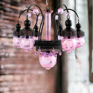 May include: A decorative chandelier with a black metal frame and six pink glass globes. The globes have a floral design and are accented with pink crystal beads. The chandelier is suspended from a chain.