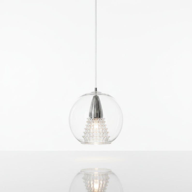 May include: A modern pendant light with a clear glass globe and a silver-colored, cone-shaped center. The light hangs from a thin white cord against a white background. The design features a textured, crystal-like element within the glass sphere.