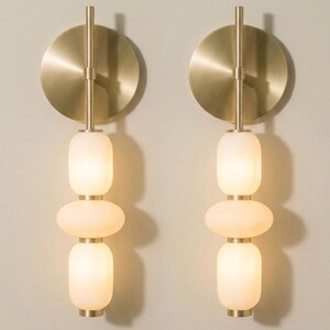 May include: Two wall sconces featuring a circular brushed brass backplate and a vertical metal rod. Each fixture holds three stacked, rounded, frosted white glass globes that emit a soft glow, creating a symmetrical lighting arrangement.