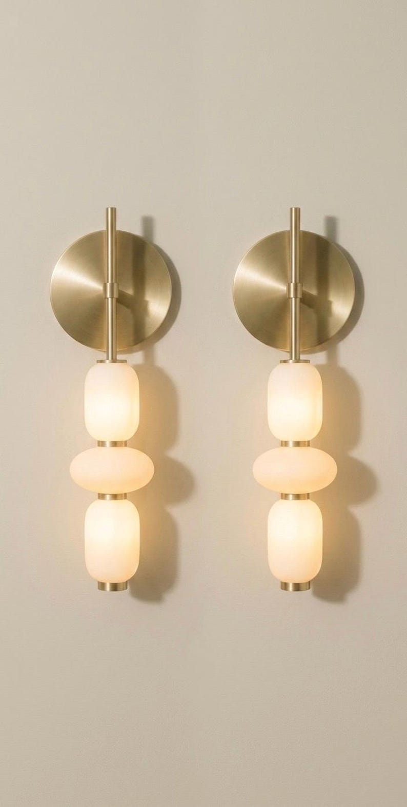 May include: Two wall sconces featuring a circular brushed brass backplate and a vertical metal rod. Each fixture holds three stacked, rounded, frosted white glass globes that emit a soft glow, creating a symmetrical lighting arrangement.