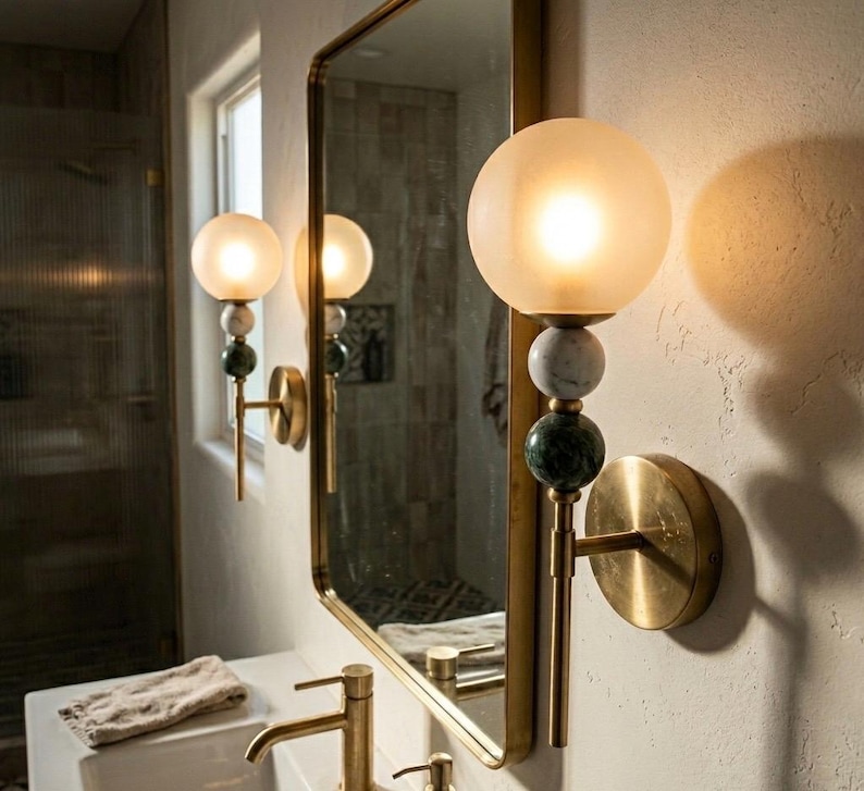 May include: A pair of wall sconces with frosted globe shades, marble accents, and brass fixtures. The sconces are mounted on a textured wall next to a mirror. The bathroom fixtures have a vintage aesthetic.