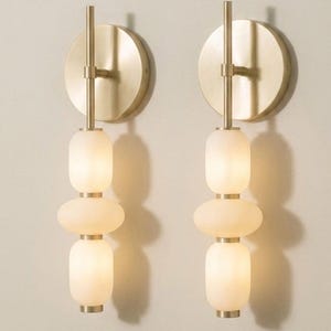 May include: A pair of wall sconces featuring a circular brushed brass backplate and a vertical rod. Each fixture holds three stacked, rounded, frosted white glass shades separated by small brass spacers, emitting a soft, warm glow.