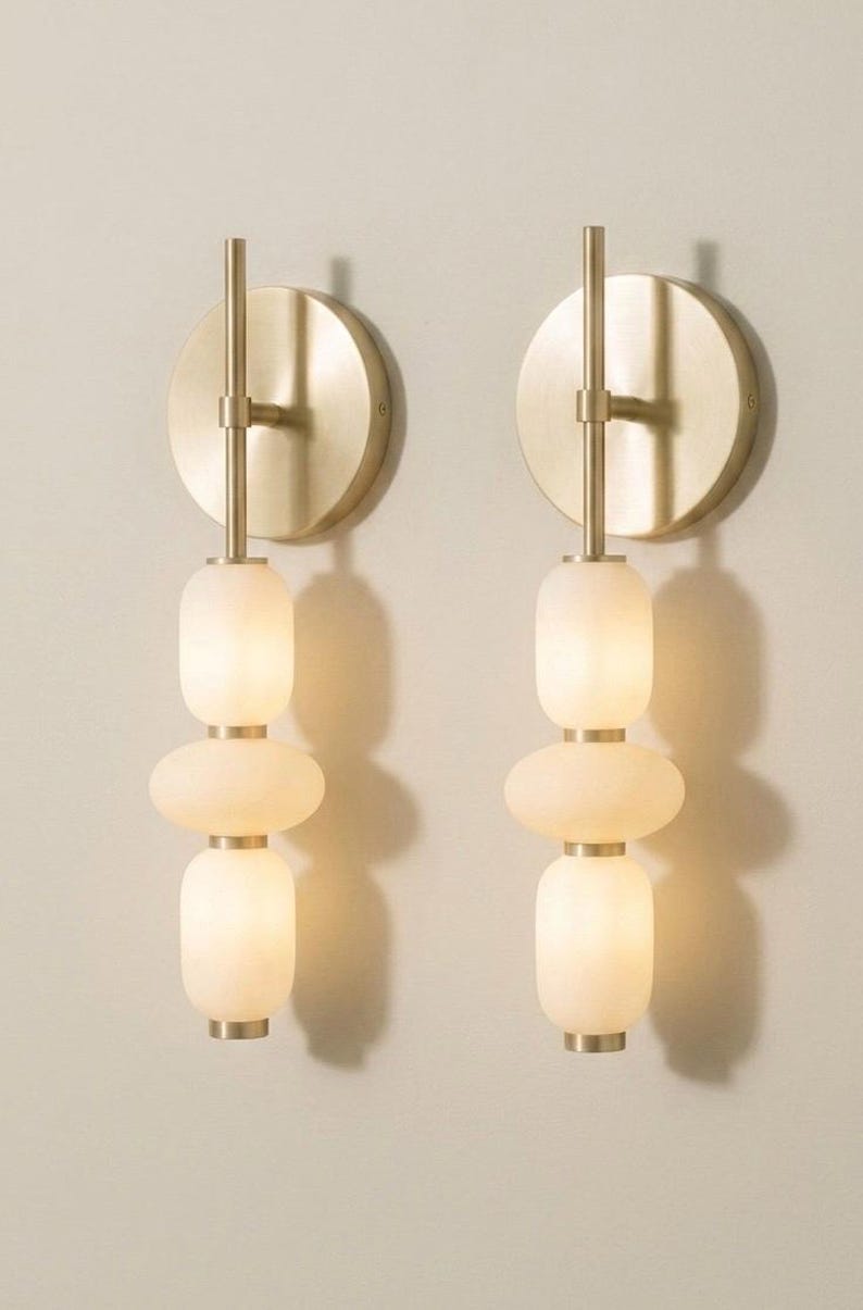 May include: A pair of wall sconces featuring a circular brushed brass backplate and a vertical rod. Each fixture holds three stacked, rounded, frosted white glass shades separated by small brass spacers, emitting a soft, warm glow.