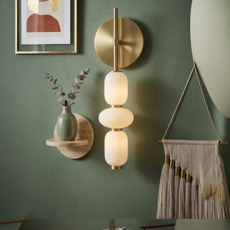 May include: A wall-mounted light fixture featuring a circular brass backplate, a vertical brass rod, and three stacked, rounded frosted glass shades. The fixture emits a soft glow, with brass accents separating each glass element.