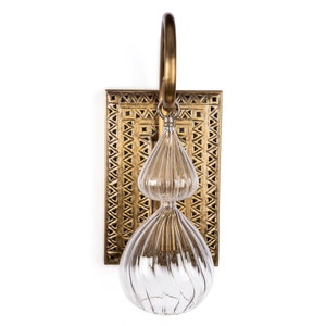 May include: A decorative wall sconce with a brass finish. The sconce features a rectangular backplate with a geometric pattern, a clear glass teardrop-shaped light fixture, and a brass loop for hanging.