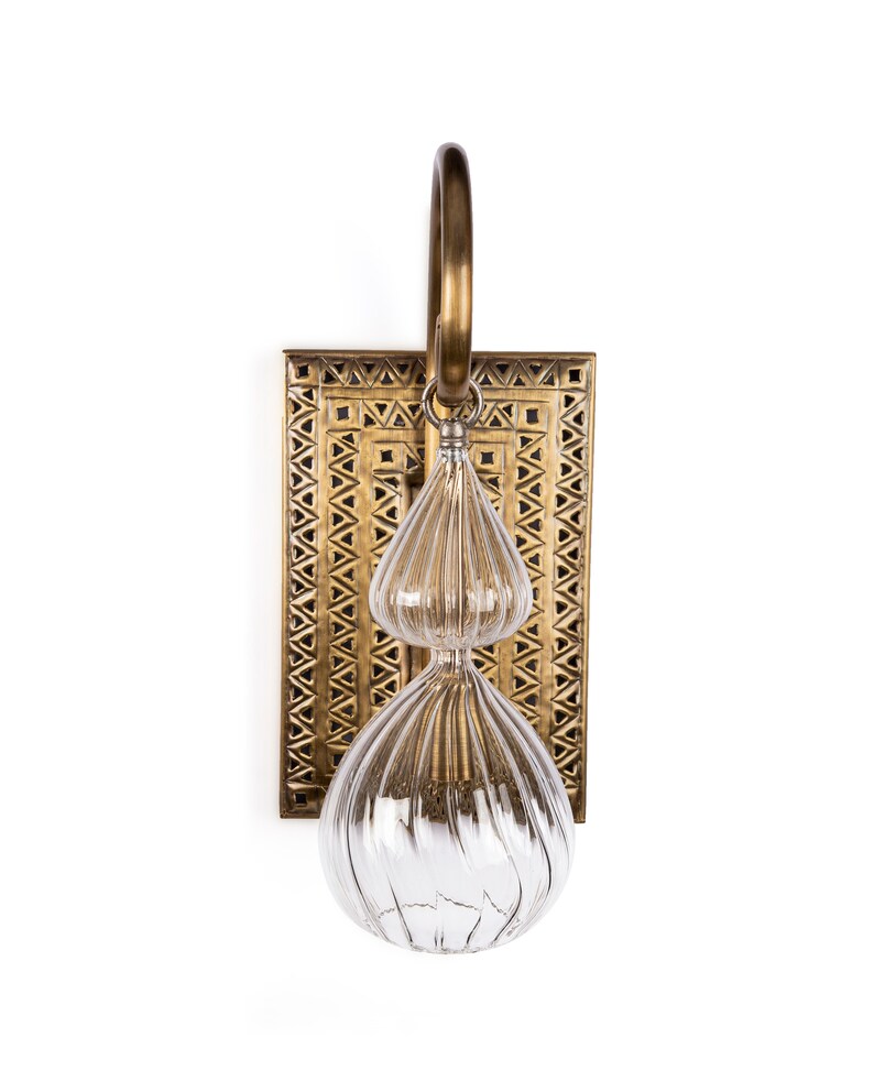 May include: A decorative wall sconce with a brass finish. The sconce features a rectangular backplate with a geometric pattern, a clear glass teardrop-shaped light fixture, and a brass loop for hanging.