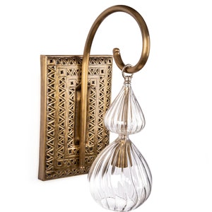 May include: A brass wall sconce with a rectangular backplate featuring a geometric pattern. A curved brass arm supports a clear, double-tiered glass shade with vertical ridges. The fixture is designed for indoor lighting.