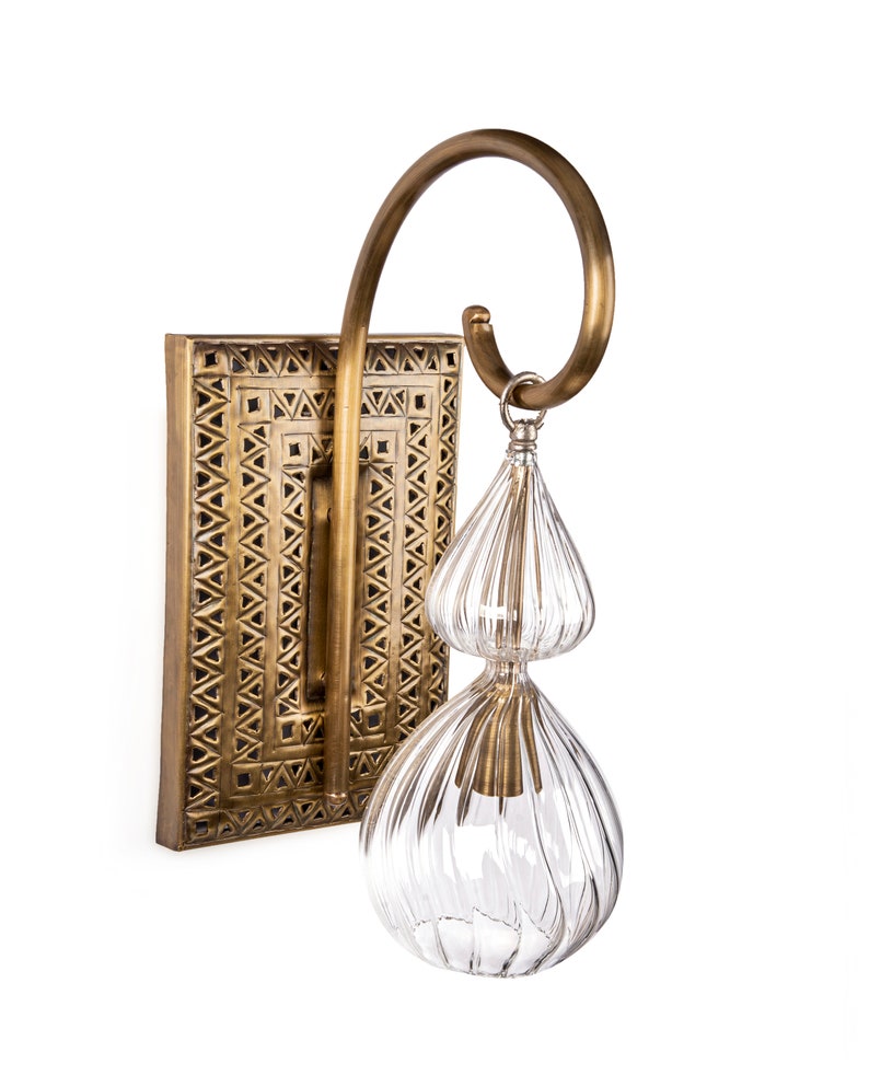 May include: A brass wall sconce with a rectangular backplate featuring a geometric pattern. A curved brass arm supports a clear, double-tiered glass shade with vertical ridges. The fixture is designed for indoor lighting.