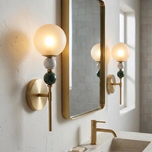 May include: A pair of wall sconces with a gold finish, featuring a frosted globe light, marble and green stone accents. The sconces are mounted on a textured white wall, with a gold-framed mirror in the background.