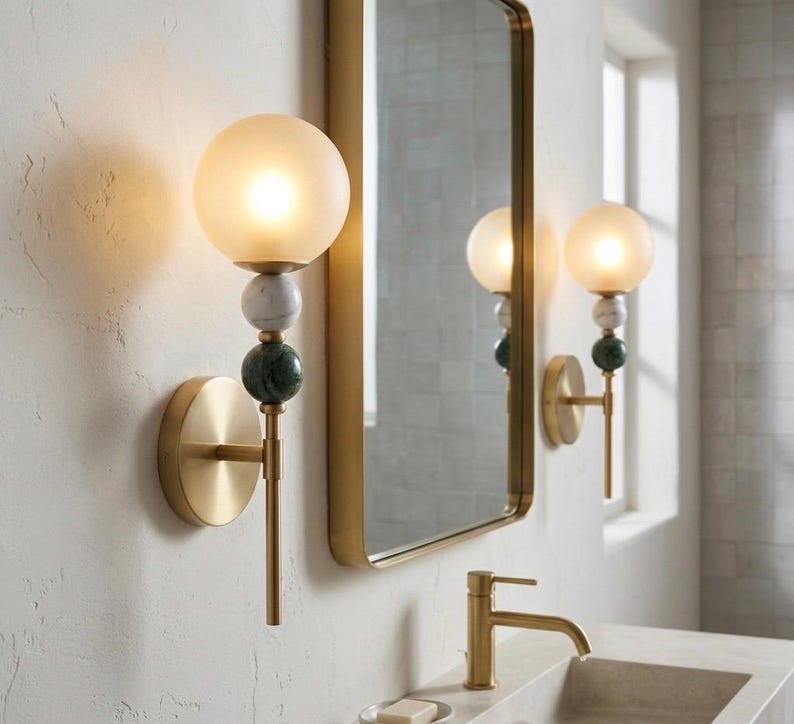 May include: A pair of wall sconces with a gold finish, featuring a frosted globe light, marble and green stone accents. The sconces are mounted on a textured white wall, with a gold-framed mirror in the background.
