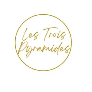 May include: A gold-colored logo featuring the words "Les Trois Pyramides" in a cursive font, enclosed within a gold circular border, set against a white background.