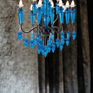May include: A vibrant blue chandelier with multiple light fixtures. The chandelier features ornate, glass-like blue accents and a dark metal frame. The bulbs are lit, casting a warm glow. The fixture hangs against a textured, neutral background.