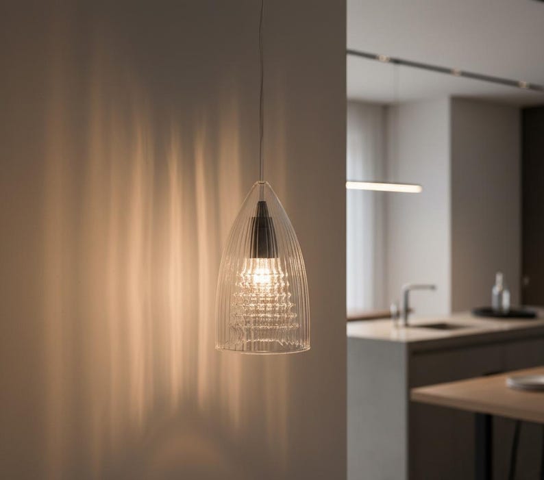 May include: A clear glass pendant light fixture with a ribbed design. The light hangs from a thin wire and illuminates a modern kitchen. The fixture has a black top and a bright light bulb.