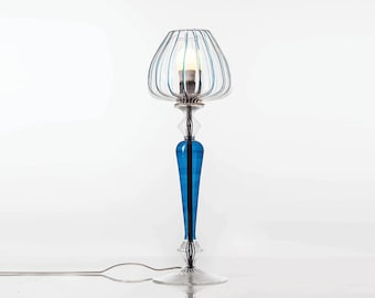 May include: A decorative table lamp with a clear glass base and a vibrant blue stem. The lamp features a cage-like shade with blue accents and a light bulb. The design is elegant and modern, suitable for home decor.