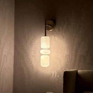 May include: A wall-mounted light fixture featuring a metallic circular base and a vertical stem supporting three stacked, rounded, translucent white glass shades that emit a soft, warm glow against the wall surface.