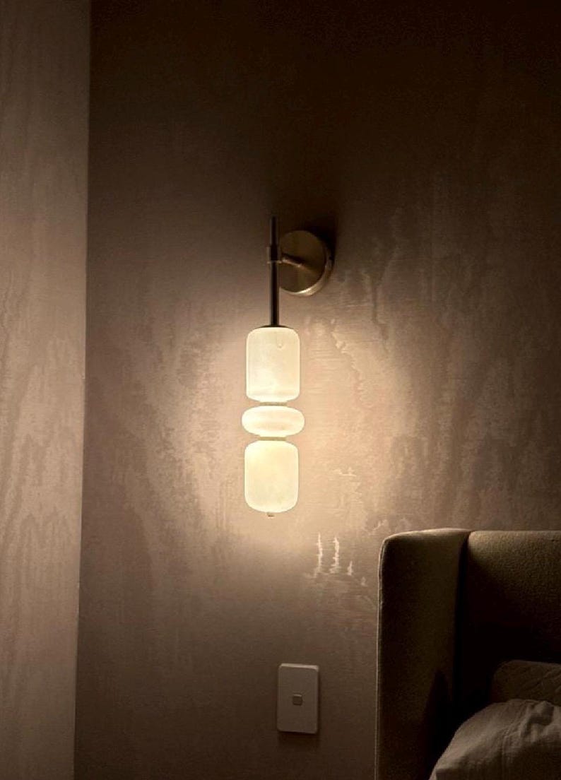 May include: A wall-mounted light fixture featuring a metallic circular base and a vertical stem supporting three stacked, rounded, translucent white glass shades that emit a soft, warm glow against the wall surface.