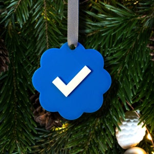 May include: A blue Christmas bauble shaped like a cloud with a white tick inside. The bauble is hanging from a grey ribbon on a Christmas tree.