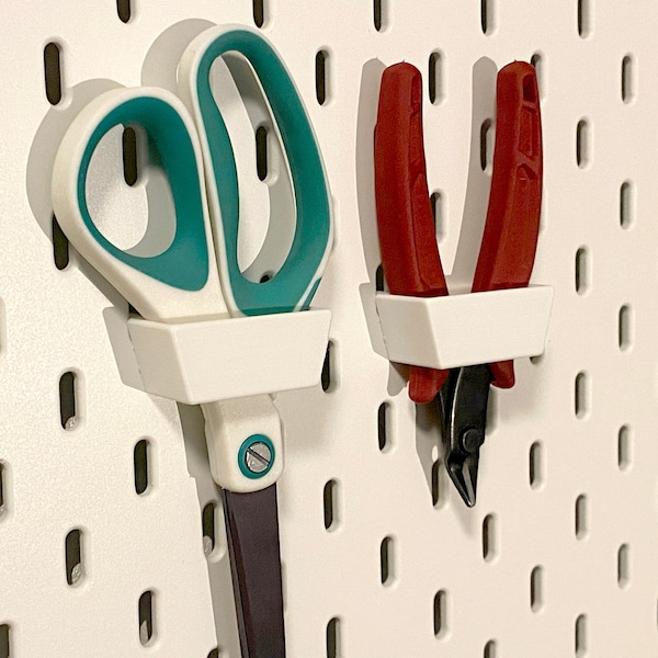 May include: White pegboard with two tool holders. The left holder has a pair of teal and white scissors. The right holder has a pair of red pliers with black handles.