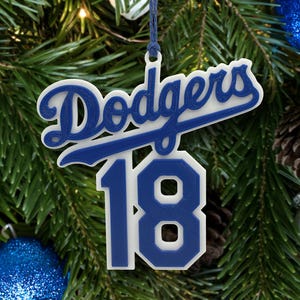 Könnte beinhalten: Blau-weißer Dodgers-Weihnachtsbaumschmuck mit der Nummer 18. Der Schmuck hängt vor einem Hintergrund aus Tannennadeln, Tannenzapfen und blauen und silbernen Kugeln. Der Teamname wird in einer markanten Schriftart angezeigt.