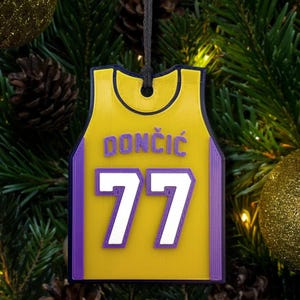 May include: A yellow and purple basketball jersey ornament with the name "DONCIC" and the number "77" in white. The ornament is hanging from a black string against a background of Christmas tree branches and ornaments.