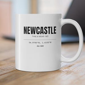 May include: White ceramic mug with the word "NEWCASTLE" in bold black letters. Below the word are the words "TYNE & WEAR - NET", coordinates, and the year "Est. 1205". The mug is on a wooden surface.
