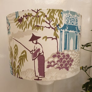May include: A white lampshade with a beige background and a colorful Asian-inspired design. The design features a person fishing in a garden with a pagoda in the background. The lampshade is on a white lamp base.
