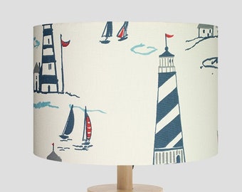 May include: A wooden table lamp with a white drum shade featuring a nautical design with blue and red sailboats and lighthouses.