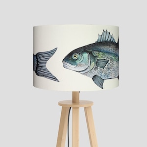 May include: A wooden table lamp with a white drum shade featuring a watercolor illustration of two fish. The fish are facing opposite directions, with one fish's tail visible on the left side of the shade and the other fish's head and body visible on the right side of the shade.