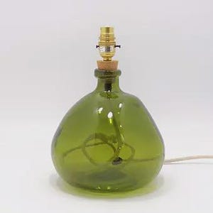 May include: A table lamp base crafted from a rounded, translucent olive green glass bottle. The neck is fitted with a cork stopper and a metallic gold-toned light bulb socket. A white electrical cord is visible inside the glass and extending outward.