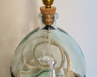 May include: A table lamp base made from a clear, light blue glass bottle with a rounded shape. A cork stopper sits in the bottle neck, supporting a brass-coloured metal lamp socket. A beige fabric-covered electrical cord is coiled inside the glass.