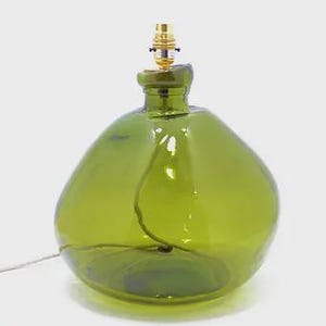 May include: A large, rounded table lamp base crafted from translucent olive green glass. The lamp features a wide, bulbous body with a narrow neck and a metallic gold-toned light socket fixture at the top. A thin electrical cord runs through the glass.