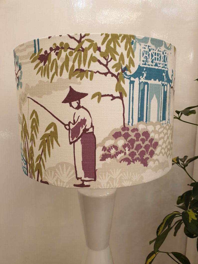 May include: A white lampshade with a beige background and a colorful Asian-inspired design. The design features a person fishing in a garden with a pagoda in the background. The lampshade is on a white lamp base.