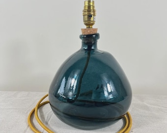 May include: A dark teal, bulbous glass lamp base with a smooth, reflective surface. The neck is fitted with a cork stopper and a metallic gold-toned light socket. A long, flexible yellow fabric-covered electrical cord is coiled around the base.
