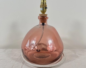 May include: A table lamp base made from a rounded, translucent dusty rose glass vessel. The neck is fitted with a cork stopper and a metallic gold-toned lamp socket fixture. A fabric-covered electrical cord runs through the centre of the glass.