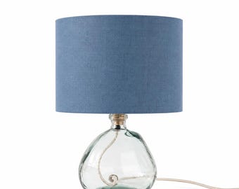 May include: A table lamp featuring a cylindrical blue fabric lampshade and a clear, rounded glass base. A white electrical cord enters the top of the glass base, with a portion of the cord coiled inside the transparent, bulbous glass structure.