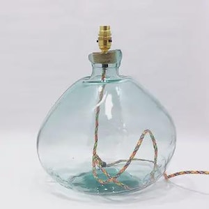 May include: A clear, light blue glass lamp base with a rounded, bulbous shape. The top features a brass-colored metal light socket fitting. A multi-colored, braided fabric electrical cord runs through the center of the glass body and exits.