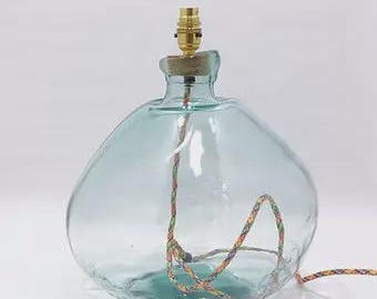 May include: A clear, light blue glass lamp base with a rounded, bulbous shape. The top features a brass-coloured metal light socket fitting. A multi-coloured, braided fabric electrical cord runs through the centre of the glass body and exits.
