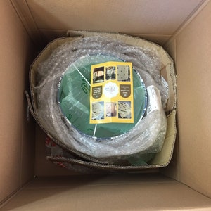 May include: A cardboard box with a round, green and blue patterned lampshade inside. The lampshade is wrapped in bubble wrap and a small card with the text "Garden Retreat" is visible.