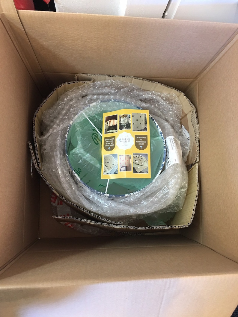 May include: A cardboard box with a round, green and blue patterned lampshade inside. The lampshade is wrapped in bubble wrap and a small card with the text "Garden Retreat" is visible.