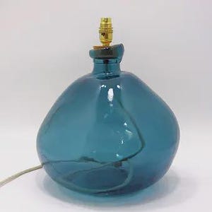 May include: A rounded, translucent teal blue glass lamp base with an irregular, organic shape. The top features a gold-colored metal light socket fitting. A thin, light-colored electrical cord extends from the side of the glass base.