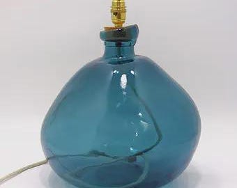 May include: A rounded, translucent teal blue glass lamp base with an irregular, organic shape. The top features a gold-colored metal light socket fitting. A thin, light-colored electrical cord extends from the side of the glass base.