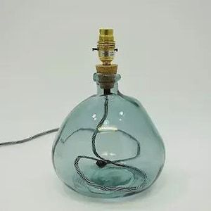 May include: A table lamp base crafted from a rounded, translucent blue-green glass bottle. The neck is fitted with a cork stopper and a metallic gold-toned light bulb socket. A black and white twisted fabric electrical cord runs through the center.