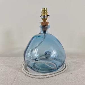 May include: A table lamp base crafted from a rounded, translucent blue glass bottle. The neck is fitted with a cork stopper and a metallic gold-toned light bulb socket. A light-colored fabric-covered electrical cord is coiled inside the glass base.