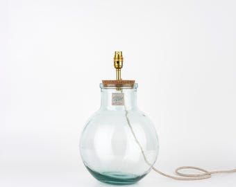 May include: A clear glass table lamp base with a round, bulbous body and a narrow neck. A cork stopper sits in the opening, topped with a gold-coloured metal lamp socket. A small rectangular label with the text USM is attached to the glass neck.