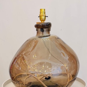 May include: A brown glass lamp with a rounded, organic shape. The lamp has a gold-colored bulb socket and a cork stopper. A beige cord runs through the lamp. The lamp is sitting on a white surface.