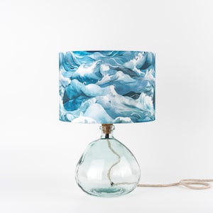 May include: A table lamp featuring a clear, organic-shaped glass base and a cylindrical lampshade decorated with a blue and white watercolour-style pattern of crashing ocean waves. A light-coloured rope cord extends from the base of the lamp.