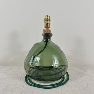 May include: A table lamp base crafted from a rounded, translucent olive green glass vessel. It features a cork stopper at the neck, a metallic brass-colored light socket, and a dark green fabric-covered electrical cord coiled inside the glass.
