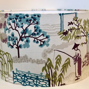 May include: A white lampshade with a blue, green, and purple floral pattern. The pattern features trees, a bridge, and a person fishing.